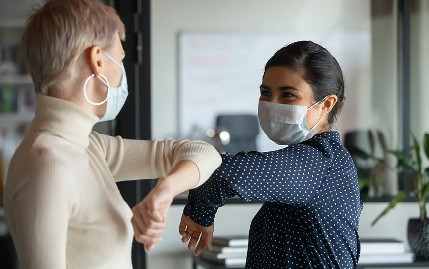 Two women wearing face masks greet each other by bumping elbows